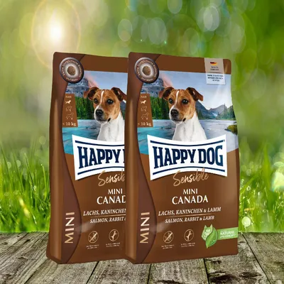 Two bags of Happy Dog Sensible MINI Canada dog food are displayed on a wooden surface with a blurred green background.