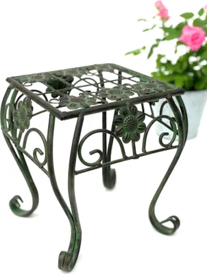 A green metal plant stand with floral details is shown against a white background, with a blurred pink flower in a pot in the background.