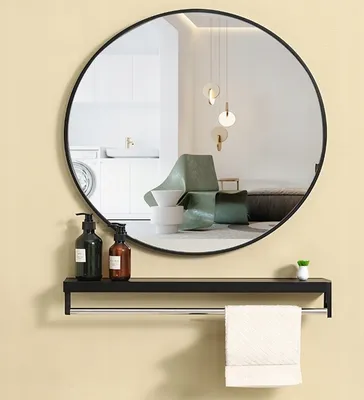 A round black mirror hangs on a light yellow wall above a black shelf with two soap dispensers, a small plant, and a towel hanging from a bar.
