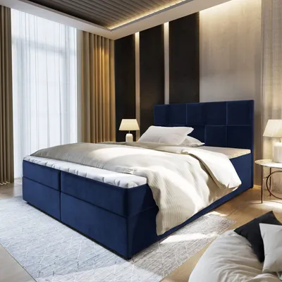 A bedroom scene with a large blue bed as the focal point, featuring a mattress and topper. The room has modern decor with textured walls, curtains, and lighting.