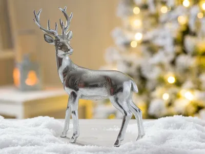 A silver deer figurine stands in artificial snow with a blurred Christmas tree and lantern in the background.