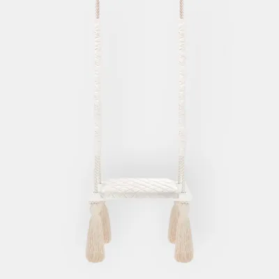 A white indoor swing with beige tassels hanging from the bottom, set against a plain white background.
