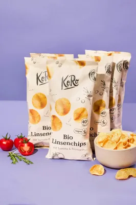 Several bags of lentil chips are arranged with fresh tomatoes and rosemary on a purple background.