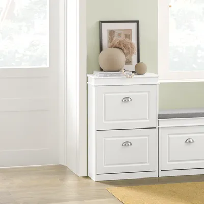A white shoe cabinet with two drawers is shown next to a window with a gray cushion on top.