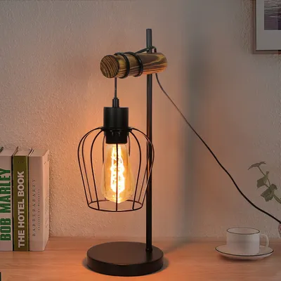 A retro-style desk lamp with a black metal cage and a wooden accent sits on a wooden desk next to some books and a cup.