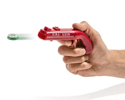 A hand holding a red bottle opener shaped like a gun, with a bottle cap in the air in front of it.