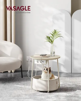 A cat sits in a fabric basket under a round side table that has a power strip and charging ports.