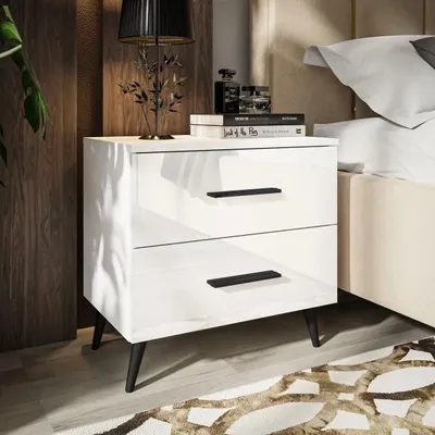 A white high-gloss nightstand with black legs and black drawer pulls, placed next to a bed. The nightstand has two drawers and is decorated with books and a lamp on top.