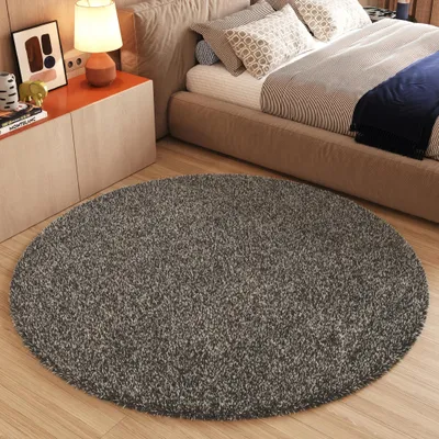 A round gray rug in a bedroom setting, placed in front of a bed and a nightstand with a lamp and artwork.