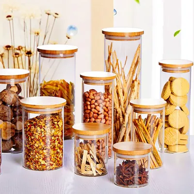 A collection of glass storage jars with bamboo lids, filled with various dry goods like pasta, nuts, crackers, and spices, arranged on a white surface.