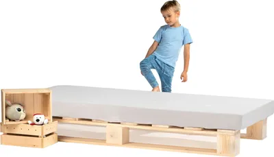 A young boy stands on a bed frame made of wooden pallets, with a mattress on top. A small wooden crate with stuffed animals sits next to the bed.