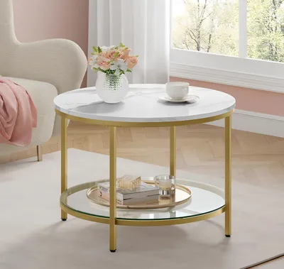 A round coffee table with a marble-like top and gold legs, placed in a living room setting. There is a vase with flowers and a teacup on the table, and a throw blanket on a nearby chair.