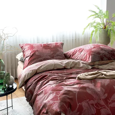 A neatly made bed with red floral patterned bedding, a pillow, and a throw blanket, next to a window with sheer curtains and a potted fern.