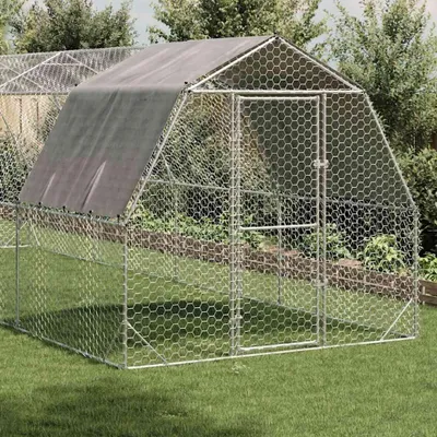 A chicken coop with a dark cover on top, set in a grassy yard with trees and bushes in the background.