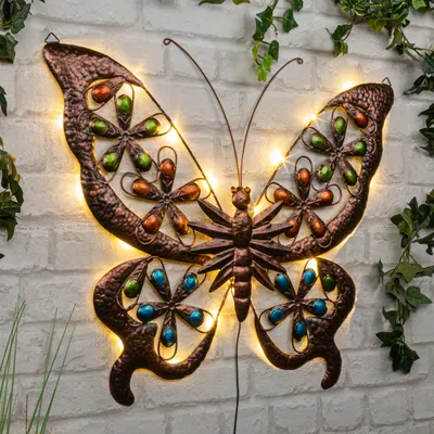 A decorative metal butterfly with LED lights is mounted on a brick wall, surrounded by greenery.
