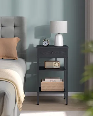 A bedside table with a lamp, alarm clock, radio, and books next to a bed.