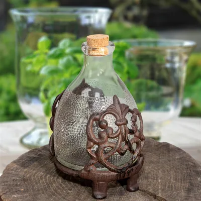 A glass bottle with a cork stopper, encased in a decorative metal stand, sits on a wooden surface. In the background, blurred glass vases hold green plants.