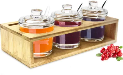 A set of three glass jars with lids and spoons, filled with different colored jams or preserves, displayed in a wooden holder. A small cluster of red berries is visible in the lower right corner.
