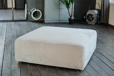 A light gray corduroy ottoman is placed on a dark wooden floor in a room with a plant and a spotlight in the background.