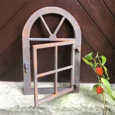 An antique-style iron window with an openable shutter, set against a wooden background, with a plant with orange fruits in the foreground.