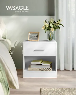 A white nightstand with a drawer and an open shelf, placed next to a bed. The nightstand has a framed picture and a vase of flowers on top, and books on the shelf. The scene is set in a bedroom with soft lighting and sheer curtains in the background.