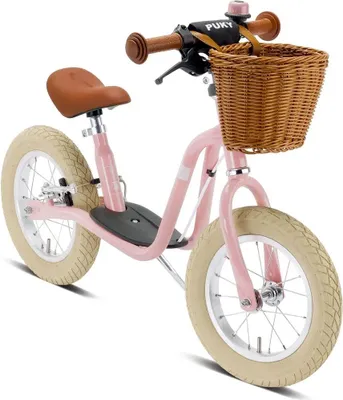 A pink balance bike with a brown seat and handlebars, and a wicker basket on the front.
