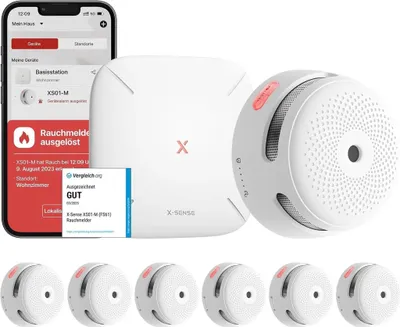 A product shot featuring a smartphone displaying a smart home app, a central base station, a smoke detector, and several additional smoke detectors arranged below.