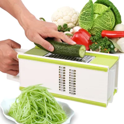 A hand is using a multi-functional grater to shred a cucumber, with a pile of shredded cucumber in a bowl in the foreground and various vegetables in the background.