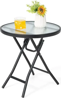 A small round glass-top table with a black metal folding frame, a glass of yellow liquid, and a vase with flowers on top.