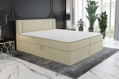 A beige boxspring bed with a mattress and topper is shown in a bedroom setting with plants and decorative elements.
