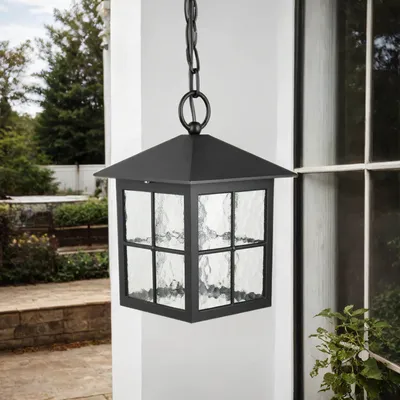A black outdoor pendant light with textured glass panels hangs from a chain against a white wall and a window.