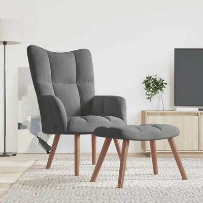 A dark gray velvet armchair and ottoman are placed on a rug in a living room setting, with a TV and cabinet in the background.