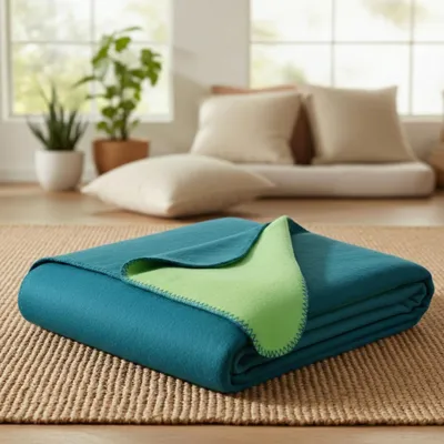 A folded teal and lime green blanket is placed on a woven rug in a living room setting with plants and cushions in the background.