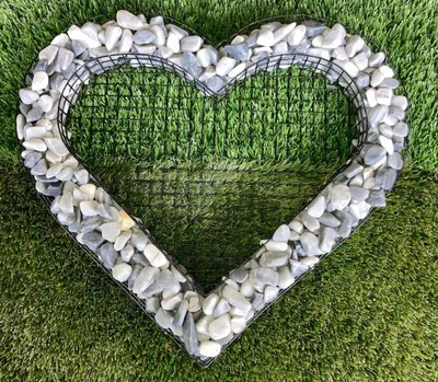 A heart-shaped wire frame filled with grey and white stones, placed on artificial grass.
