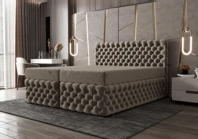 A luxurious bedroom scene featuring a large, tufted boxspring bed with a matching headboard and base, set against a textured wall with geometric accents and modern pendant lights. A nightstand and a desk with a chair are visible in the background.