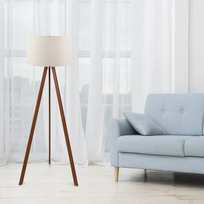 A floor lamp with a tripod base stands next to a light blue sofa in a room with sheer white curtains.