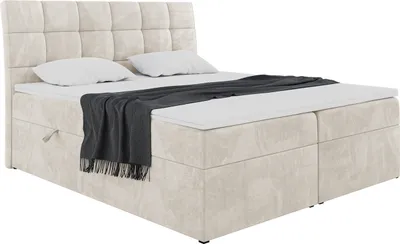 A studio shot of a cream-colored boxspring bed with a headboard, mattress, and topper. A dark gray blanket is draped over the bed.