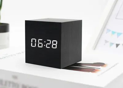A black cube-shaped digital clock displaying "06:28" is placed on a book.