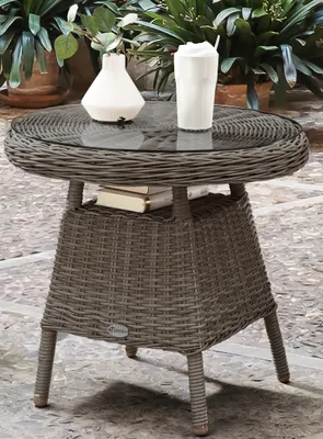 A round wicker table with a glass top, a vase, and a drink on it, placed outdoors on a stone patio with plants in the background.