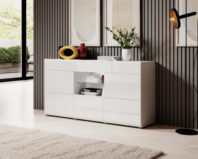 A modern white sideboard is displayed in a living room setting, with decorative items on top and a floor lamp nearby.
