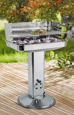 A stainless steel pedestal grill with a burger on the top rack, set up on a wooden deck outdoors with greenery in the background.