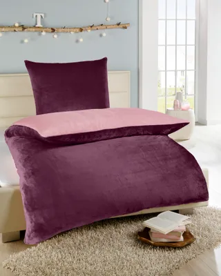 A bed with purple and pink bedding, a pillow, and a small stack of books on a rug.