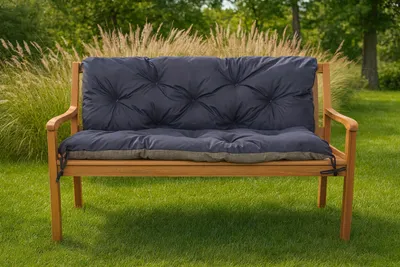 A dark blue tufted cushion is placed on a wooden garden bench, set against a backdrop of tall grasses and trees in a garden.