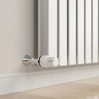 A close-up shot of a white radiator thermostat valve attached to a white radiator and a light-colored wall.