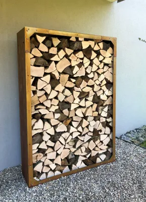 A metal firewood rack filled with chopped wood, placed outdoors on a gravel surface next to a wall.