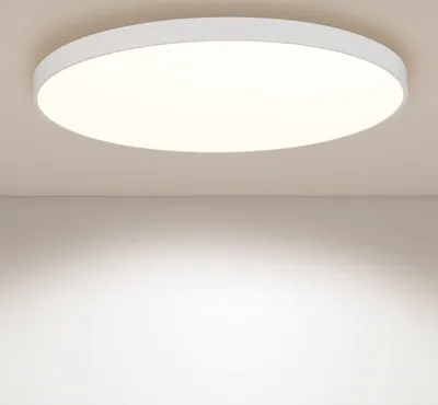 A white, round LED ceiling light is mounted on a light beige ceiling, casting a bright, warm light downwards onto a white wall.