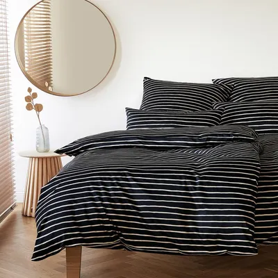 A neatly made bed with black and white striped bedding, a round mirror on the wall, and a small side table with a vase and decorative branches.