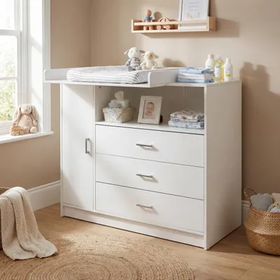 A white baby changing table with drawers and a door, set up in a nursery room with natural light coming from a window.