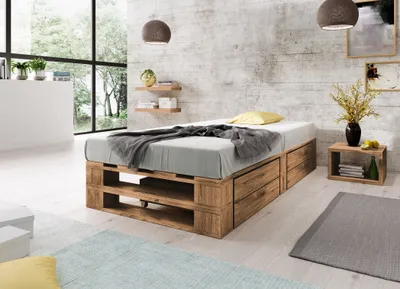 A bedroom with a wooden pallet bed frame, a mattress, and bedding, set against a textured concrete wall with large windows overlooking trees.