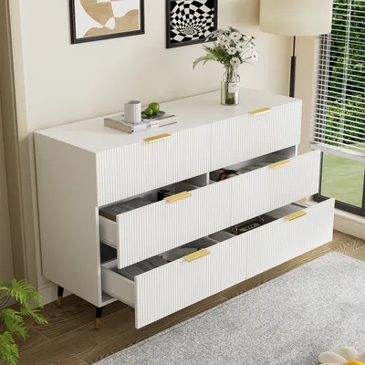 A white sideboard with six drawers, some of which are open, displaying storage compartments. The sideboard is placed on a wooden floor with a rug in front of it. Two framed pictures hang on the wall behind it, and a floor lamp is visible to the right.